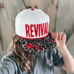 Person wearing a white cap with 'REVIVAL' printed on it, held by another person.