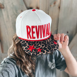 Person wearing a white cap with 'REVIVAL' printed on it, held by another person.