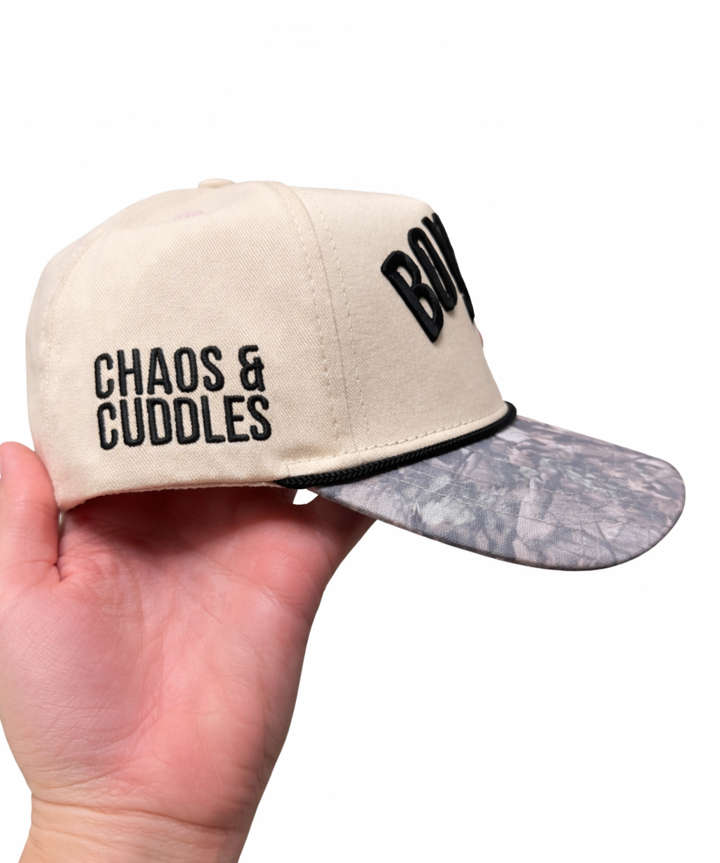 Beige cap with text 'Chaos & Cuddles' on the side held by a hand on a white background