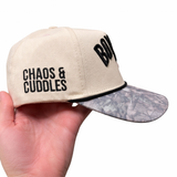 Beige cap with text 'Chaos & Cuddles' on the side held by a hand on a white background