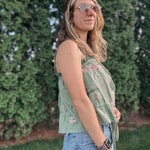 Woman wearing a green patterned top and blue jeans standing in front of green bushes.