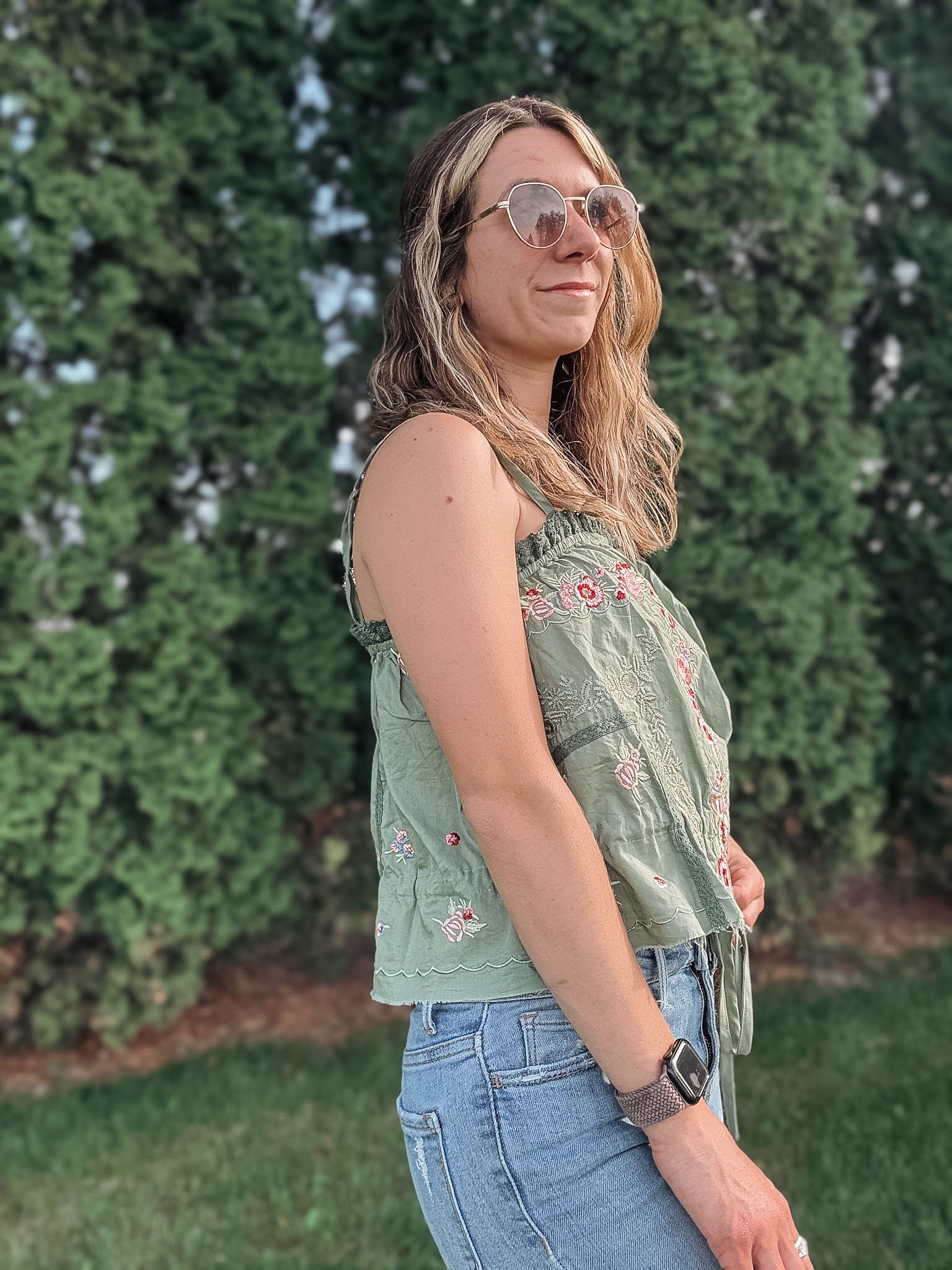 Woman wearing a green patterned top and blue jeans standing in front of green bushes.