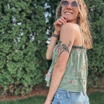 Woman wearing a green floral top and blue jeans with sunglasses, standing against a green bush.