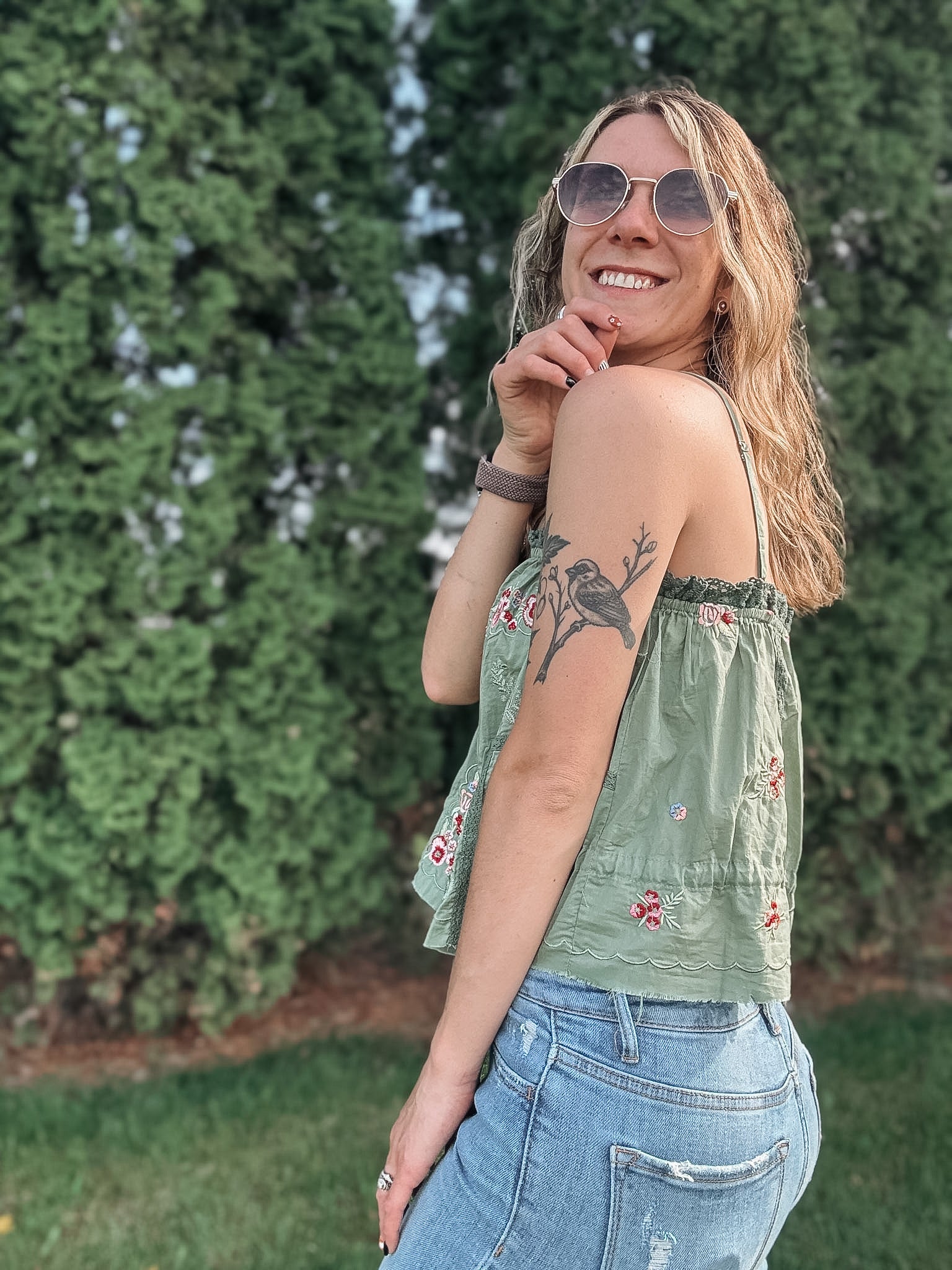 Woman wearing a green floral top and blue jeans with sunglasses, standing against a green bush.