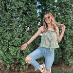 Woman in a green top and blue jeans posing outdoors with greenery in the background