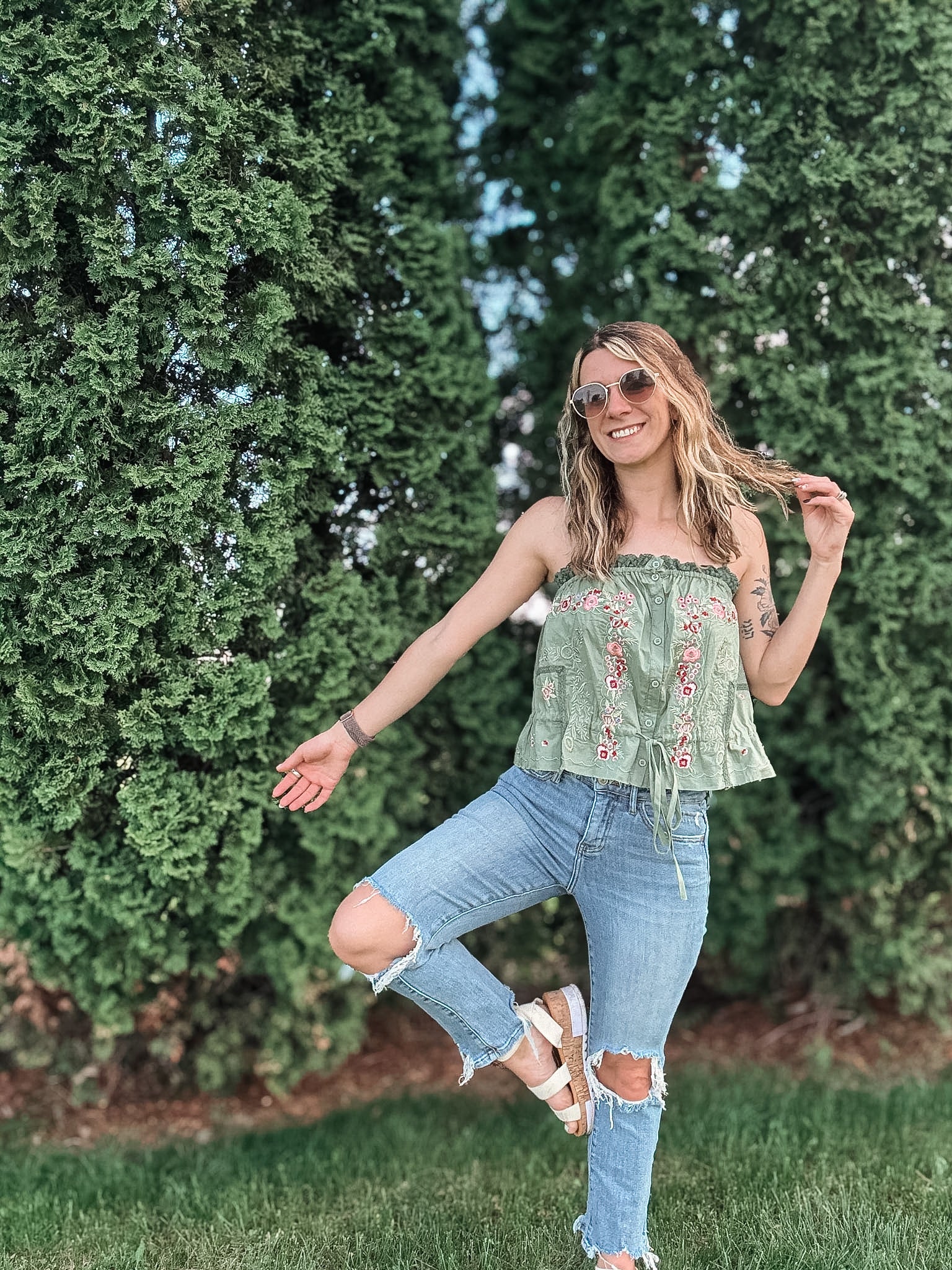 Woman in a green top and blue jeans posing outdoors with greenery in the background