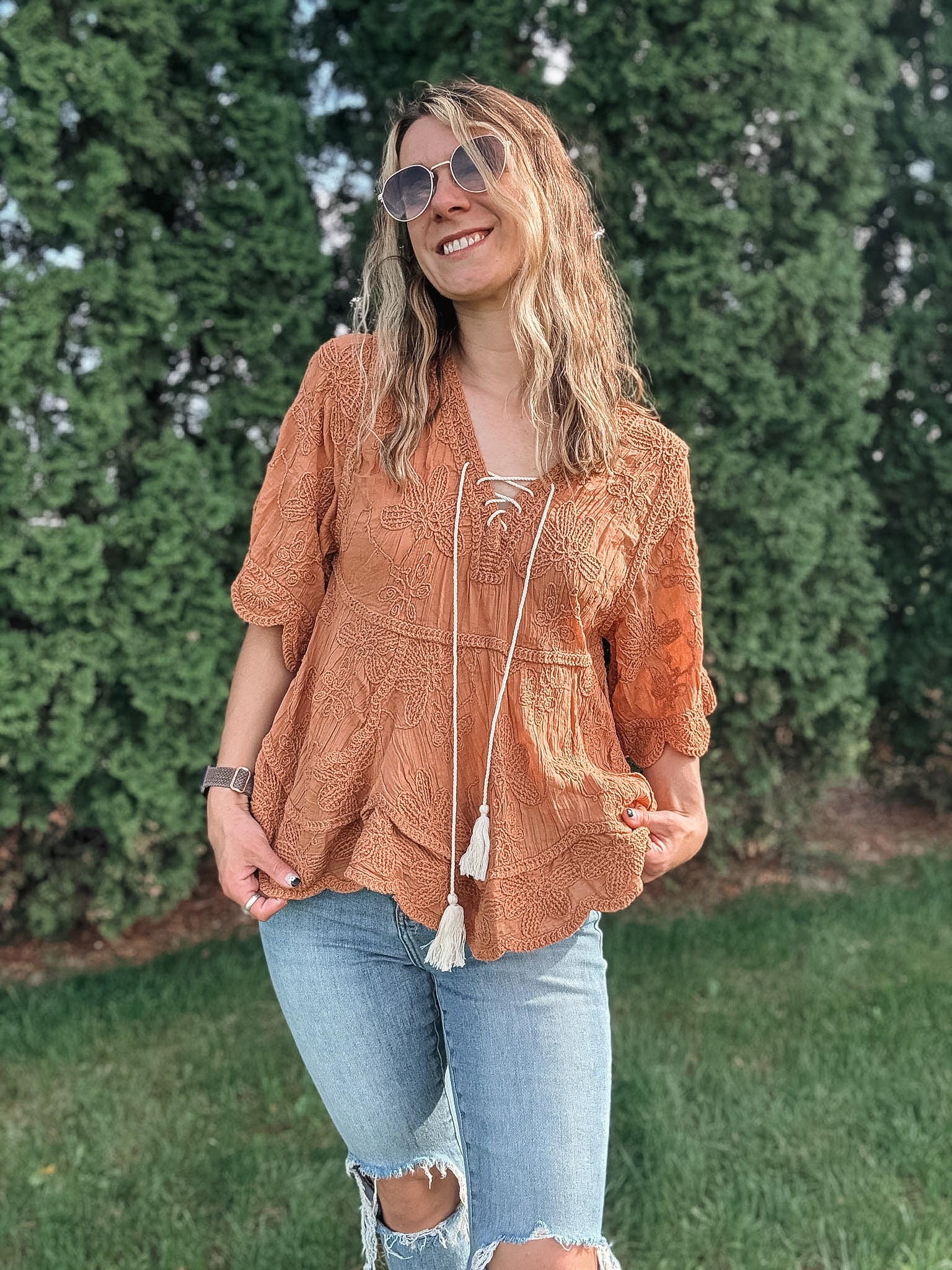 Woman wearing a brown lace top and ripped jeans standing outdoors with greenery in the background
