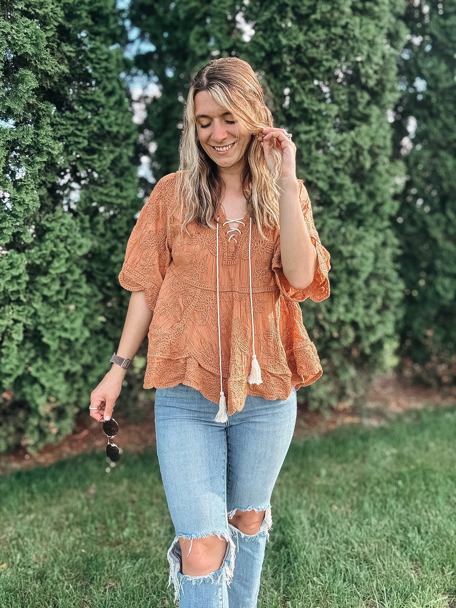 Woman wearing an orange lace top and ripped jeans standing outdoors with greenery in the background