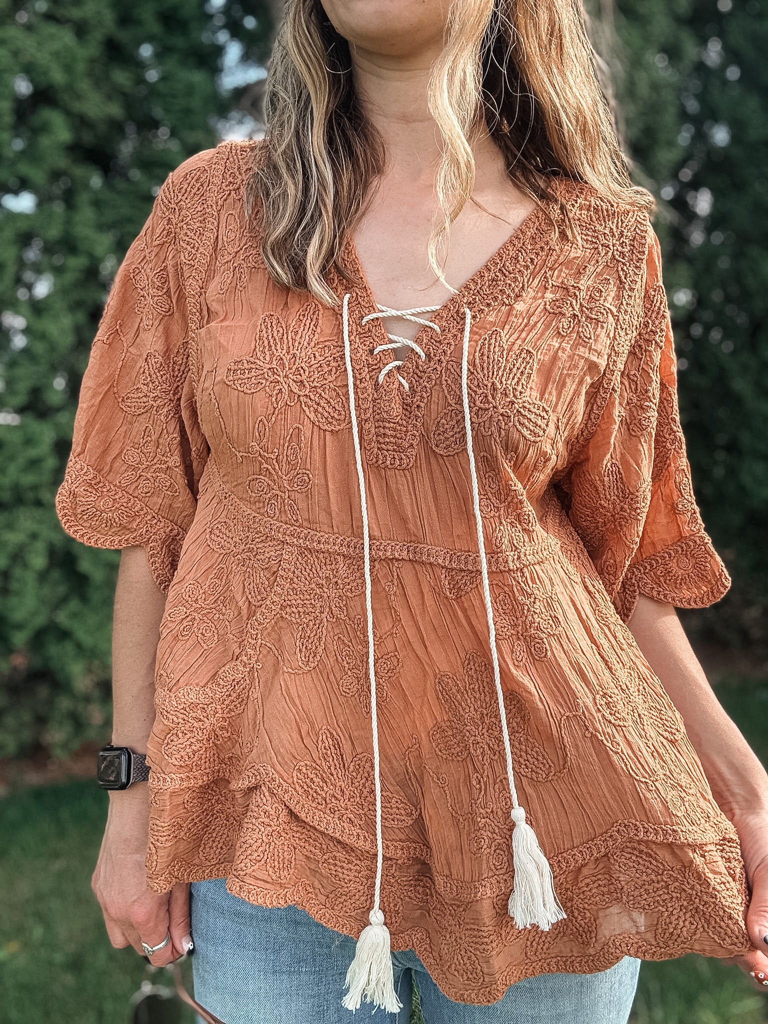 Woman wearing a brown lace-up top with tassels in front of green foliage