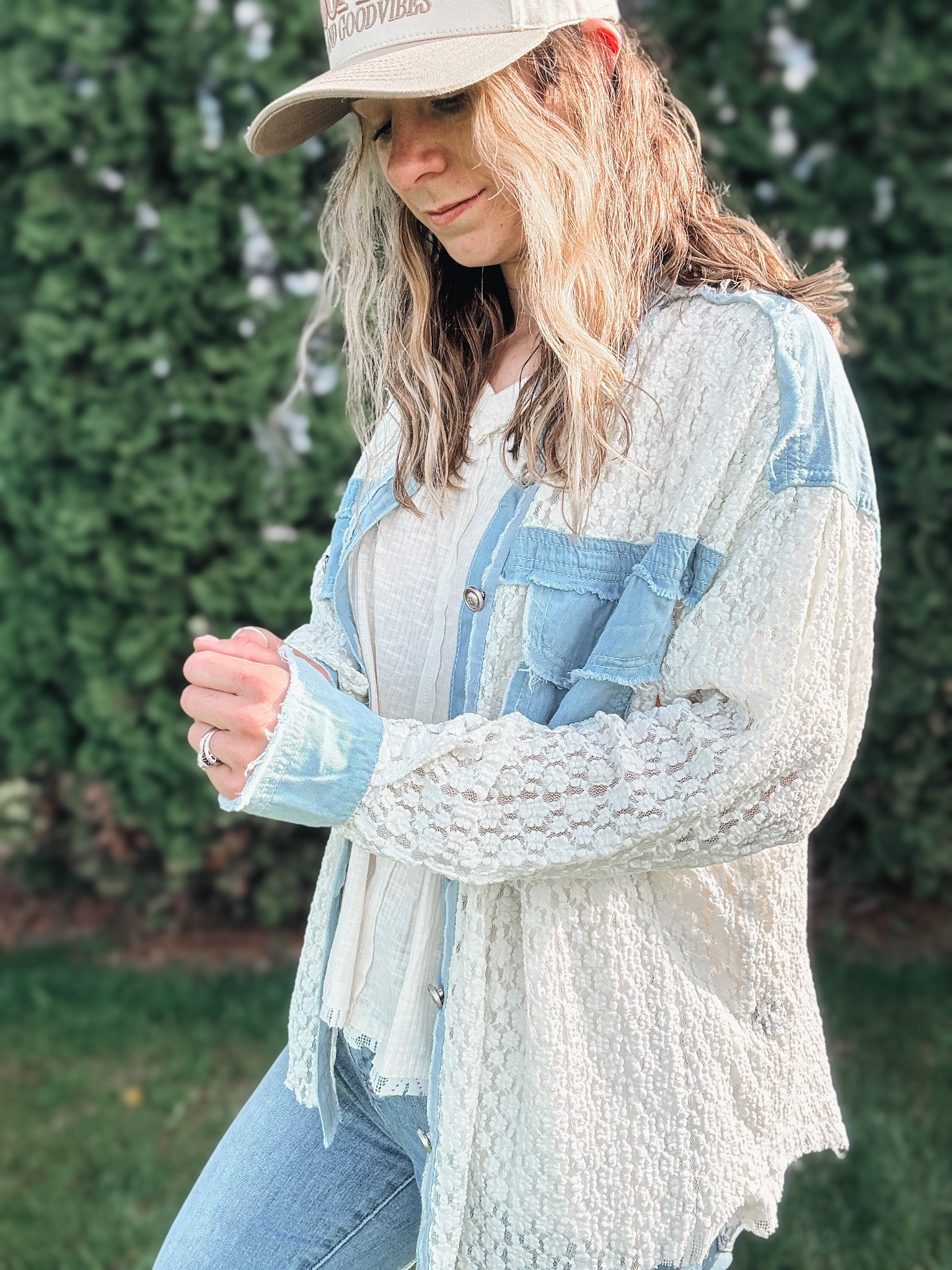 Woman wearing a light blue denim jacket over a white lace top with a beige cap outdoors.