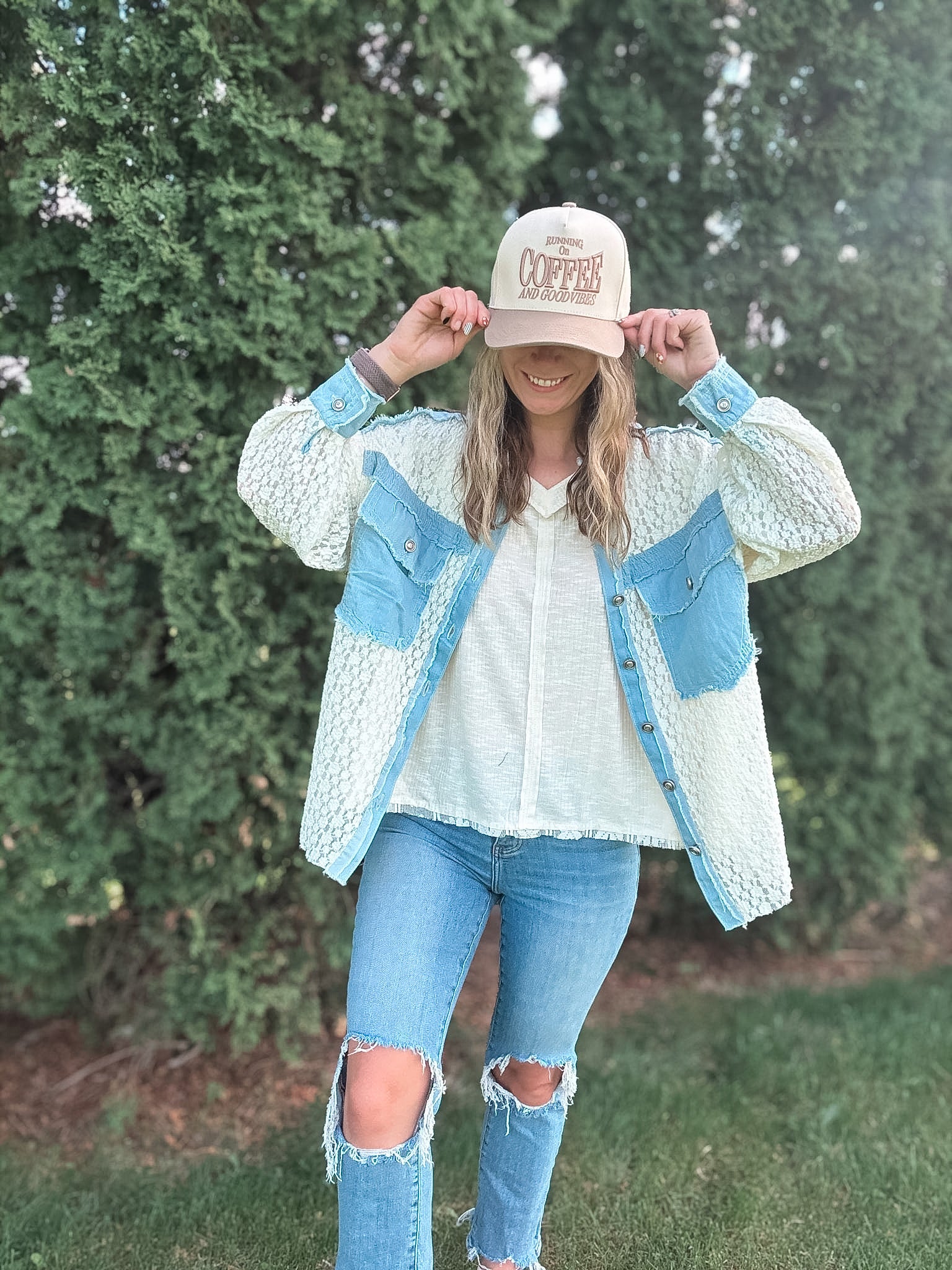Person wearing a light blue denim jacket with a white shirt and ripped jeans, standing in front of green bushes.