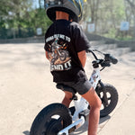 Child wearing a black t-shirt with a graphic and text, sitting on a balance bike.