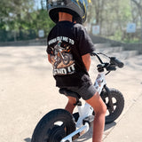 Child wearing a black t-shirt with a graphic and text, sitting on a balance bike.