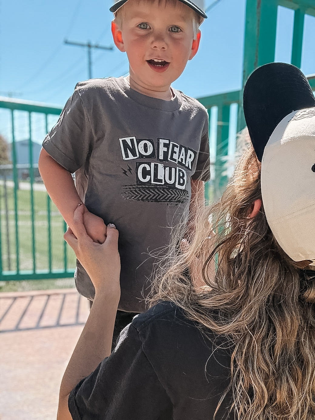 Person holding a child wearing a 'No Fear Club' shirt at a skate park.