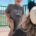 Person holding a child wearing a 'No Fear Club' shirt at a skate park.