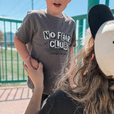 Person holding a child wearing a 'No Fear Club' shirt at a skate park.