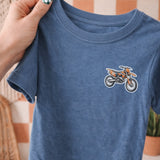 Blue t-shirt with a dirt bike graphic held by a hand against a checkered wall.