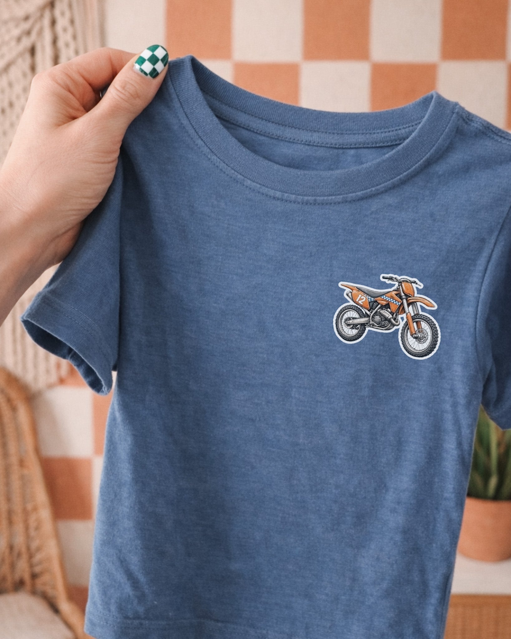 Blue t-shirt with a dirt bike graphic held by a hand against a checkered wall.