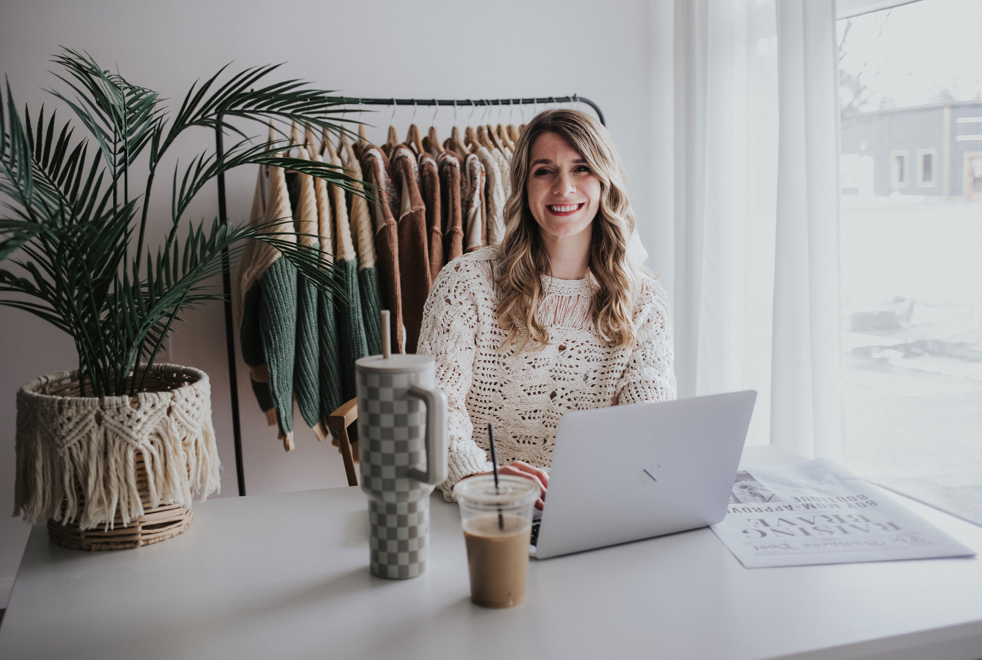Raising Brave's founder sitting in the natural light while working on her laptop with a coffee and rack of stylish women's cardigans