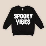 A black long-sleeve sweatshirt with 'SPOOKY VIBES' printed in a white dripping font across the chest, displayed flat against a light beige background.
