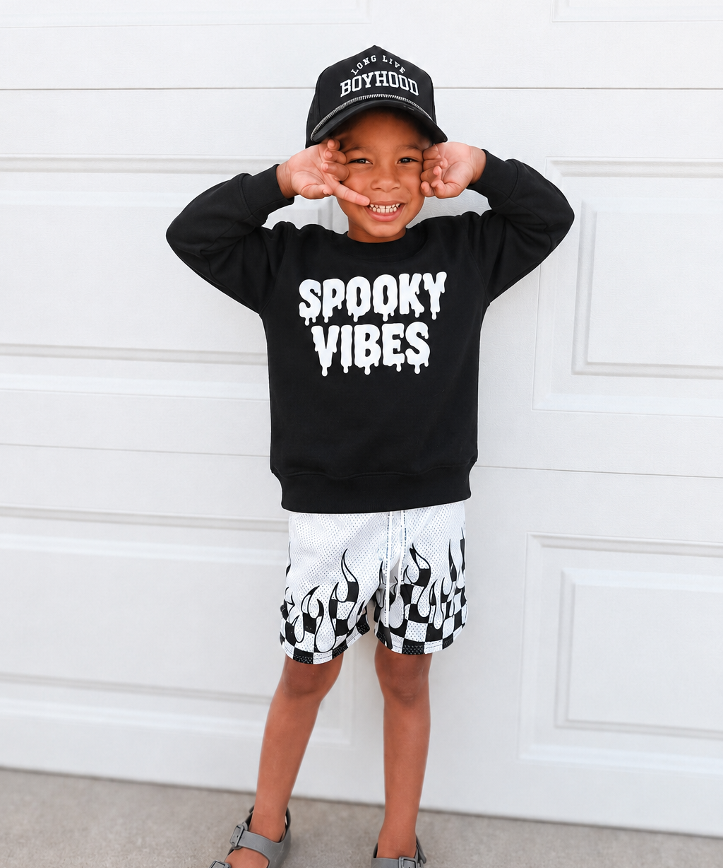 A young boy posing in front of a white garage door, wearing a black crewneck sweatshirt with 'SPOOKY VIBES' printed in white dripping letters, a black baseball cap, black and white checkered shorts, and gray sandals.