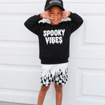A young boy posing in front of a white garage door, wearing a black crewneck sweatshirt with 'SPOOKY VIBES' printed in white dripping letters, a black baseball cap, black and white checkered shorts, and gray sandals.