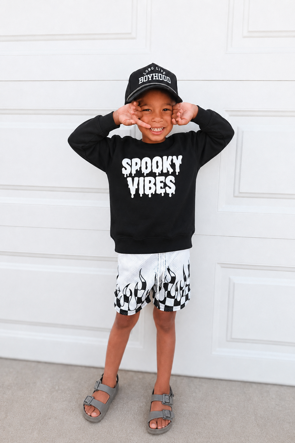 A young boy posing in front of a white garage door, wearing a black crewneck sweatshirt with 'SPOOKY VIBES' printed in white dripping letters, a black baseball cap, black and white checkered shorts, and gray sandals.